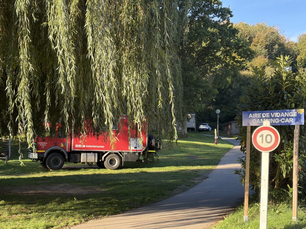 Roter Mercedes-Benz Expeditions-LKW von Niekerkensreise auf dem Flower Camping La Chênaie in Yport, Normandie. Der Wohnmobilstellplatz liegt auf einer grünen Wiese unter einer großen Trauerweide, neben der Zufahrt zur Wohnmobil-Entsorgungsstation.