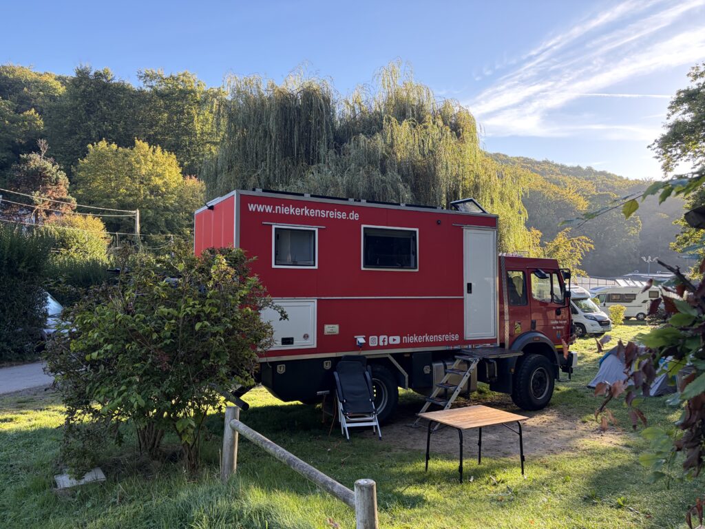 Roter Mercedes-Benz 1224 AF Expeditions-LKW von Niekerkensreise auf einem grünen Stellplatz des Flower Camping La Chênaie in Yport, Normandie. Umgeben von Bäumen und Sträuchern.