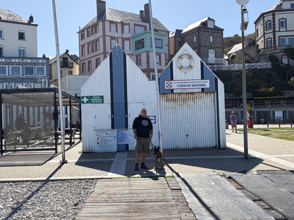 Lars von Niekerkensreise mit Malinois Elvis vor der Seenotrettungsstation SNSM am Strand von Yport in der Normandie. Im Hintergrund stehen die typischen weiß-blauen Strandgebäude und historischen Häuser des Küstenortes.