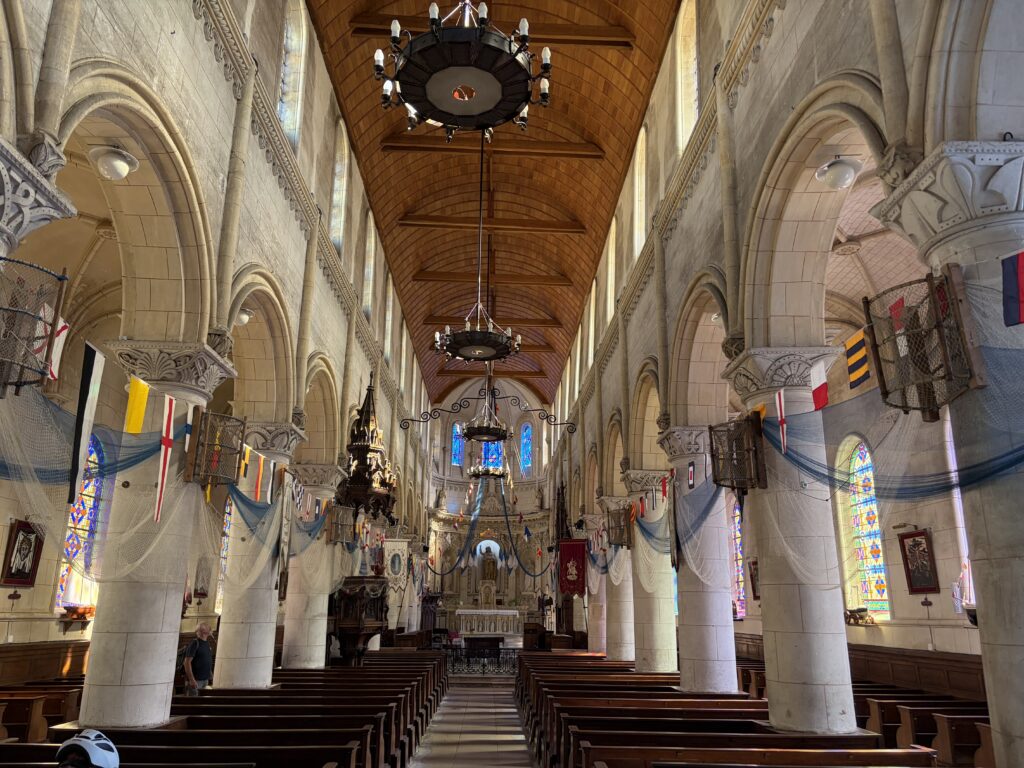 Innenraum der Kirche von Yport in der Normandie mit hohen romanischen Rundbögen, bunten Buntglasfenstern, hölzernem Tonnengewölbe und maritimen Dekorationen aus Fischernetzen und Signalflaggen.