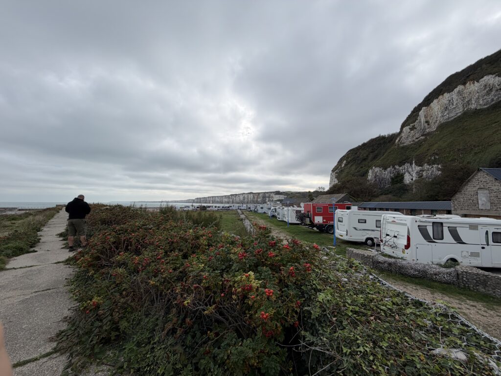 Wohnmobilstellplatz Saint-Valery-en-Caux in der Normandie mit zahlreichen Wohnmobilen, darunter ein roter Expeditions-LKW, direkt unter den weißen Kreidefelsen der Alabasterküste am Meer.