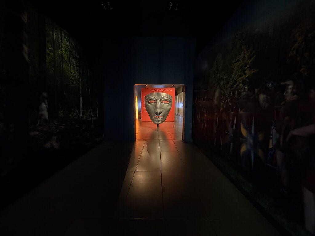 Ausstellung im Varusschlacht-Museum Kalkriese bei Bramsche mit großer römischer Gesichtsmaske vor rotem Hintergrund und beleuchtetem Durchgang.