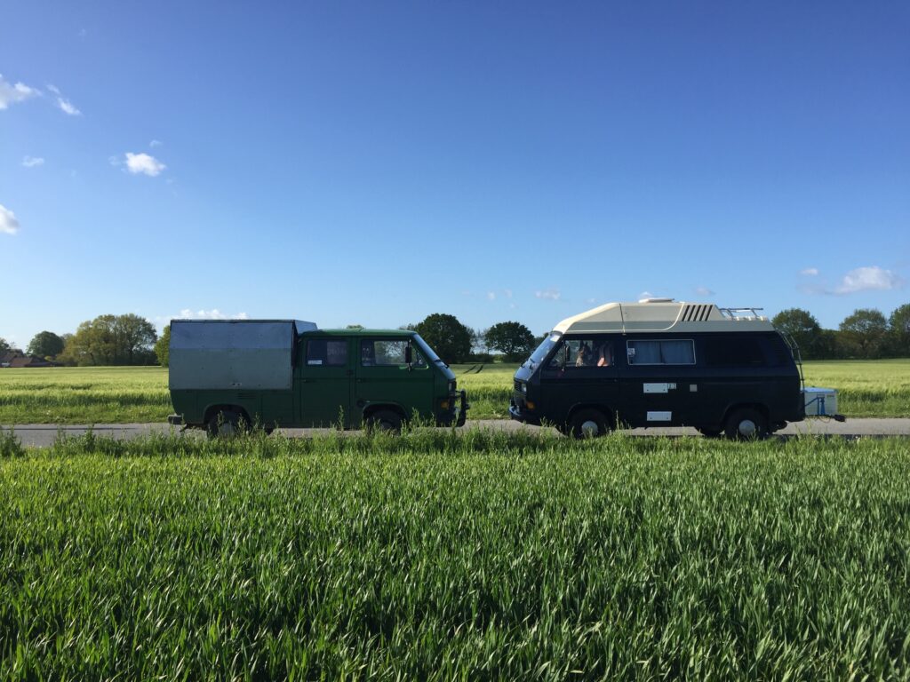 Zwei VW T3 auf einer Landstraße: Links Lars' grüne VW T3 Doka mit speziellem Aufbau für seine Arbeit als Hufschmied, rechts unser schwarzer VW T3 Weinsberg Camper „Der Doc“ mit Hochdach.
