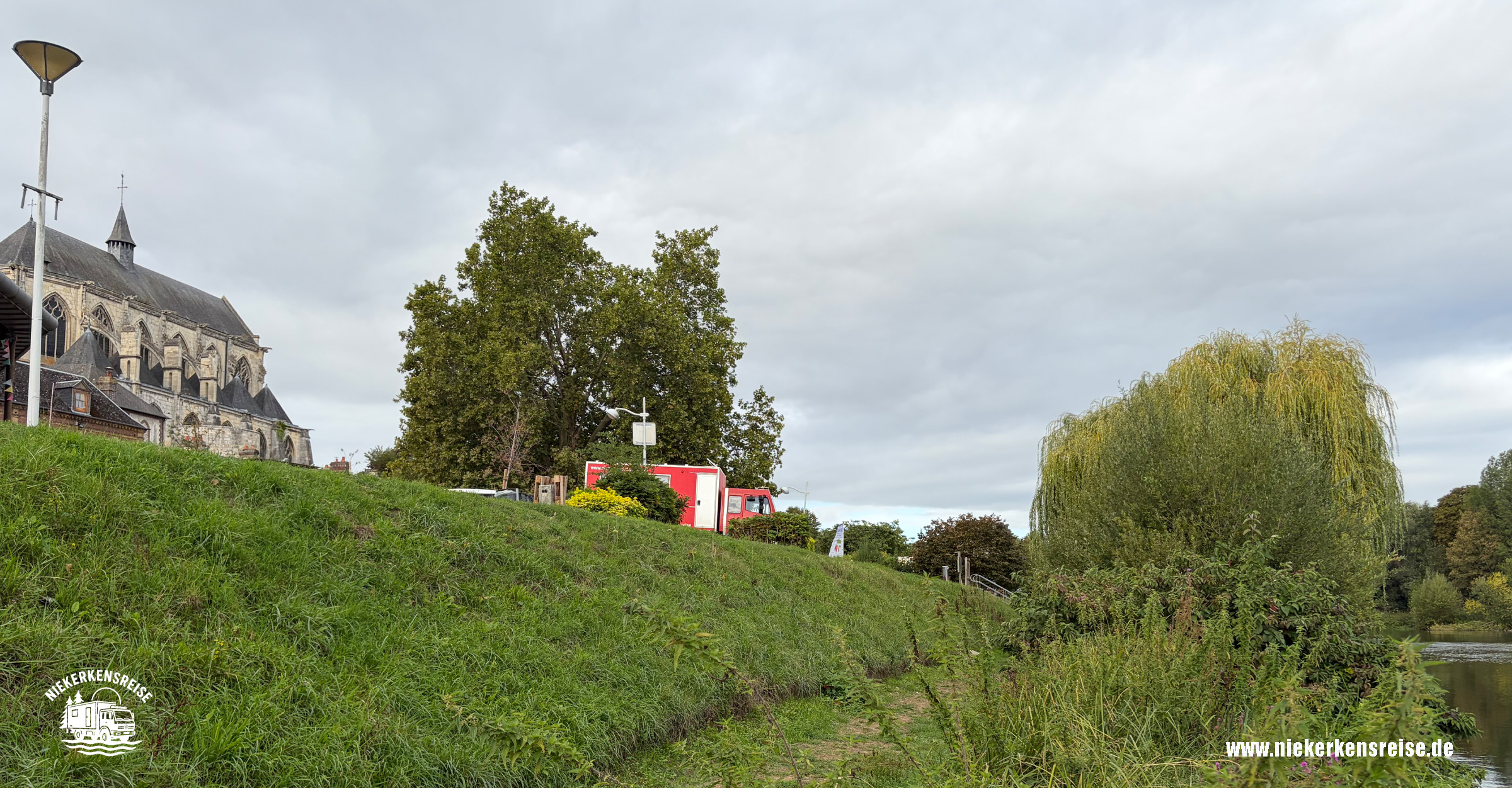 Blick vom Ufer der Eure auf den Wohnmobilstellplatz in Pont-de-l’Arche in der Normandie. Im Hintergrund stehen ein rotes Expeditionsmobil und die historische Kirche Notre-Dame-des-Arts.