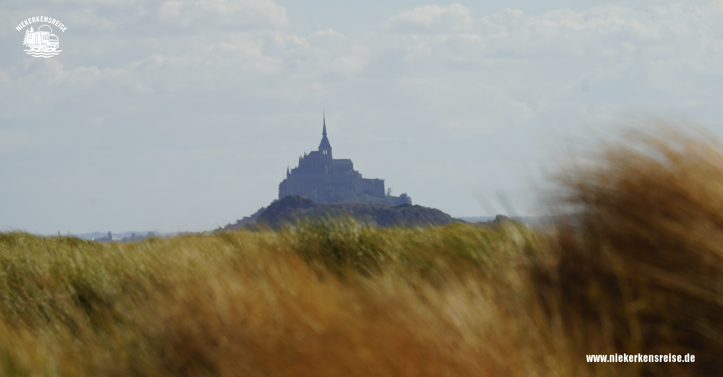Mont-Saint-Michel – Blick von den Dünen bei Dragey-Ronthon Blick von den Dünen bei Dragey-Ronthon auf den Mont-Saint-Michel in der Normandie. Im Vordergrund wiegen sich goldene Dünengräser im Wind, während die berühmte Klosterinsel am Horizont zu sehen ist.