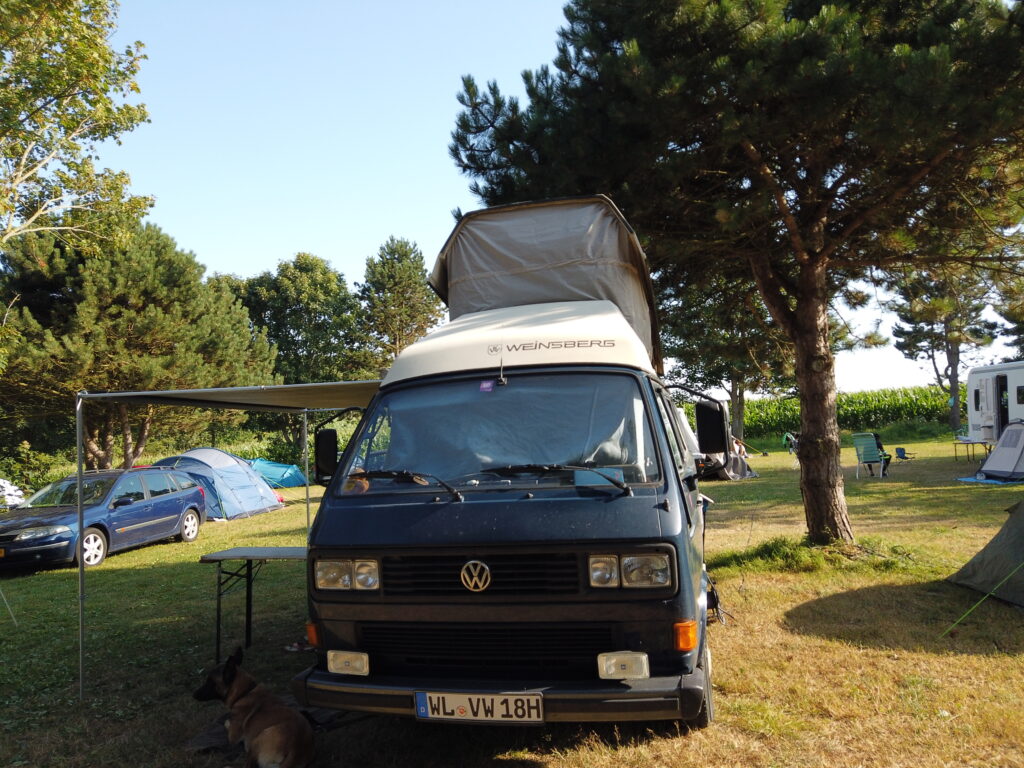 VW T3 Westfalia Weinsberg Camper mit aufgestelltem Hochdach und Markise auf dem Naturcampingplatz La Pâture in Yport, Normandie. Der Bulli steht auf einer grünen Wiese unter Bäumen.